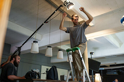 sur un plateau de tournage, un photographe sur un escabeau règle un projecteur fixé sur une girafe
